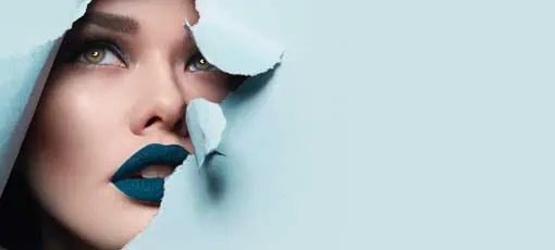 das Gesicht eines jungen schönen Mädchen mit einem hellen Make-up und puffblauen Lippen Peers in ein Loch in blauem Papier.